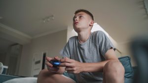 Concentrated teenager plays online video game using controller, sits on sofa